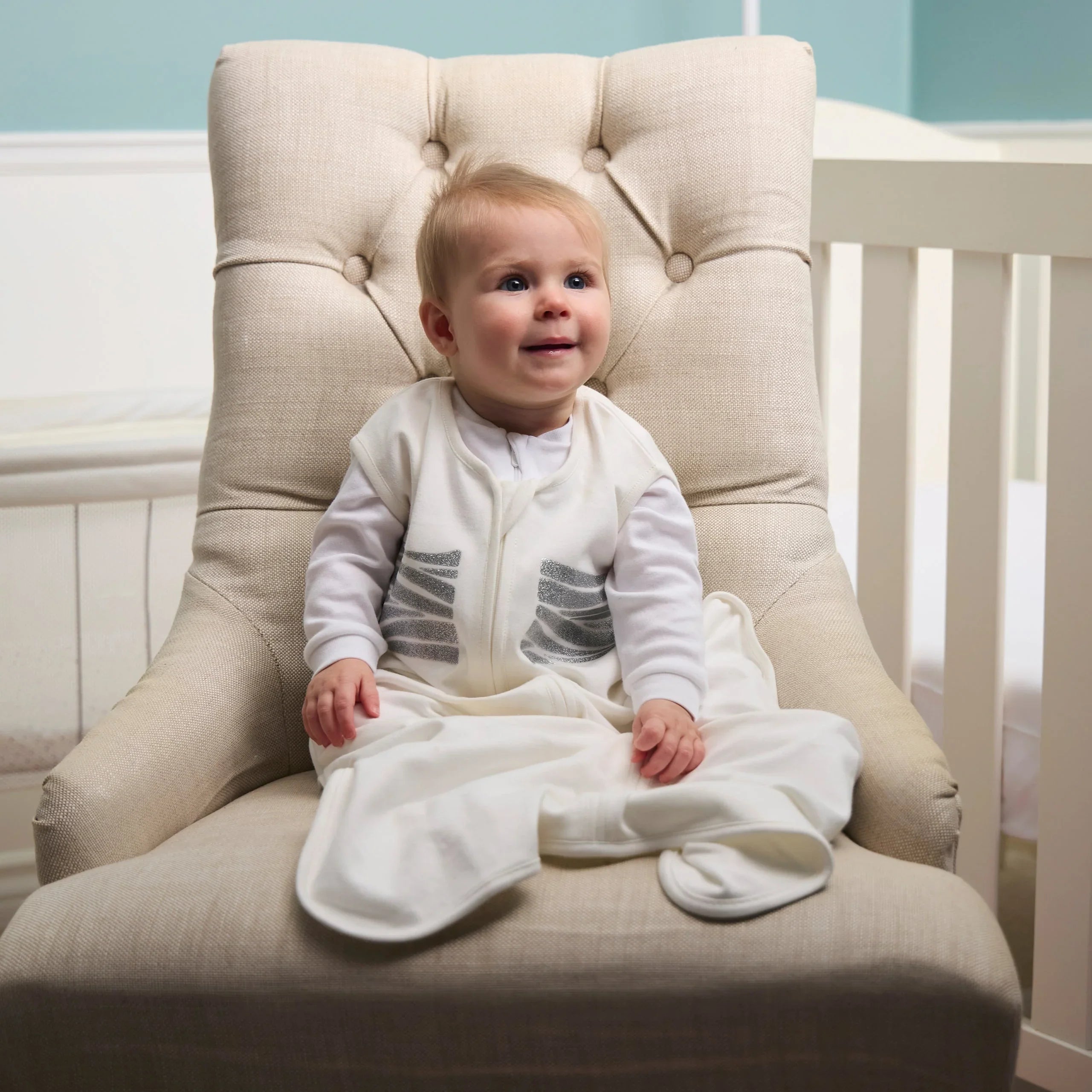 Baby in a white outfit sitting on a beige chair in a nursery.