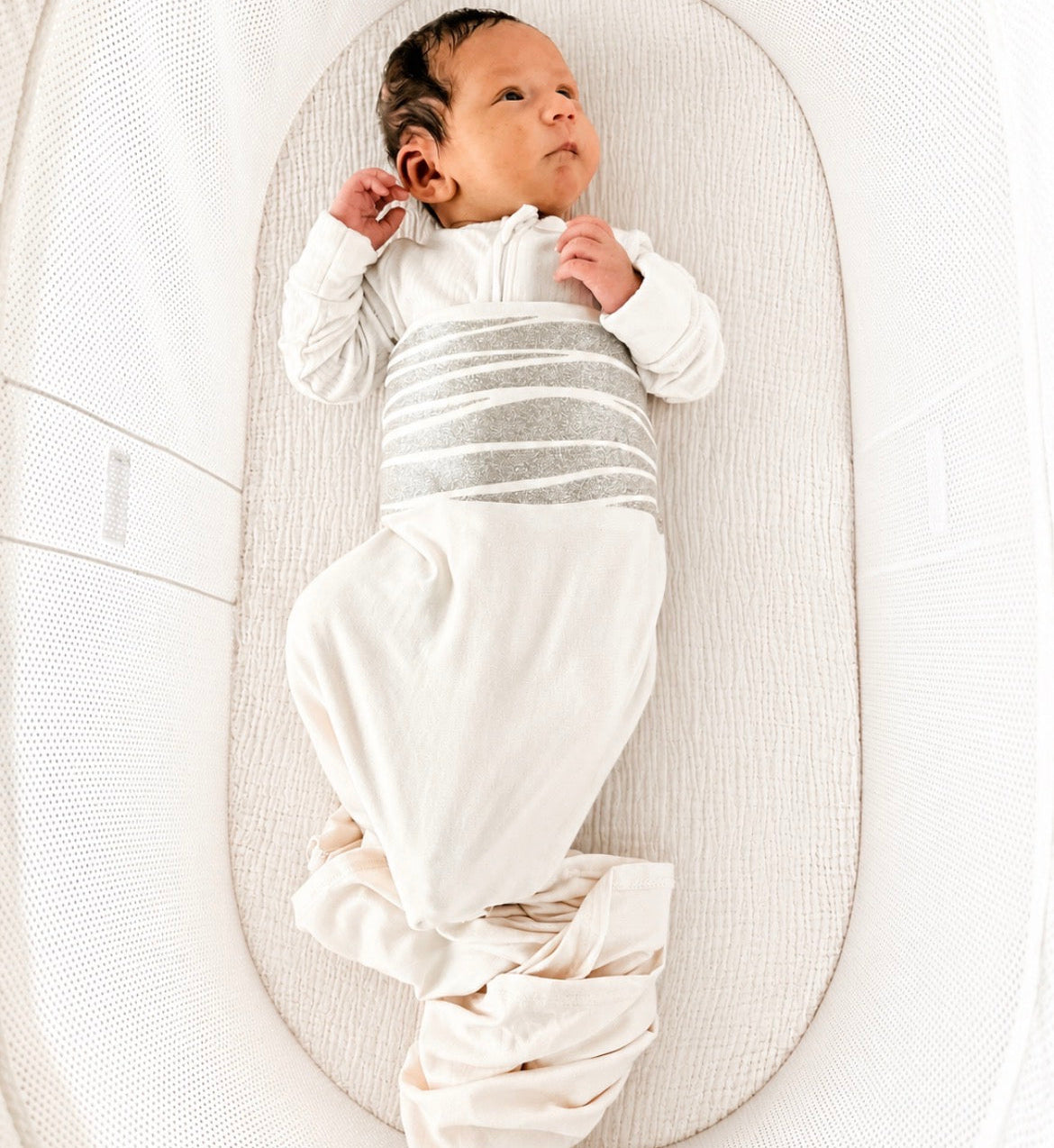 A newborn baby rests comfortably in a white bassinet, wrapped in the Baby Shower Bundle | 0 - 6 months swaddle blanket featuring gray stripes across the middle, looking up with one arm bent near their face.