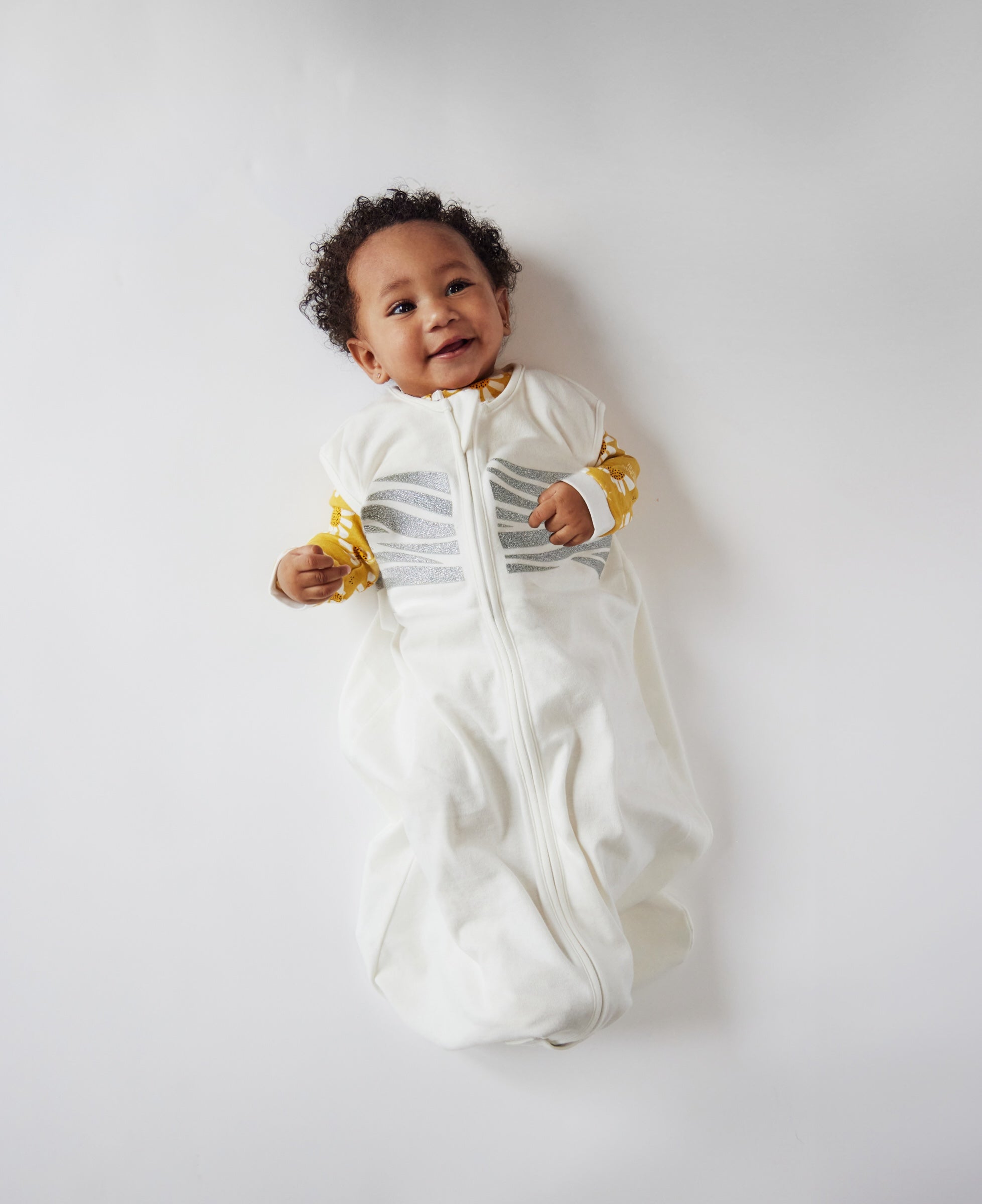 A baby wearing a white zip-up sleeping bag with gray horizontal stripes and yellow cuffs, lying on a light gray background.