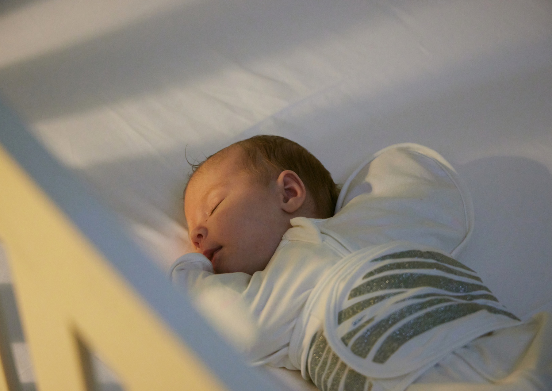 A baby sleeps peacefully on its side in a white crib, wrapped in a white swaddle with gray stripes. The scene is softly lit, creating a calm and serene atmosphere.