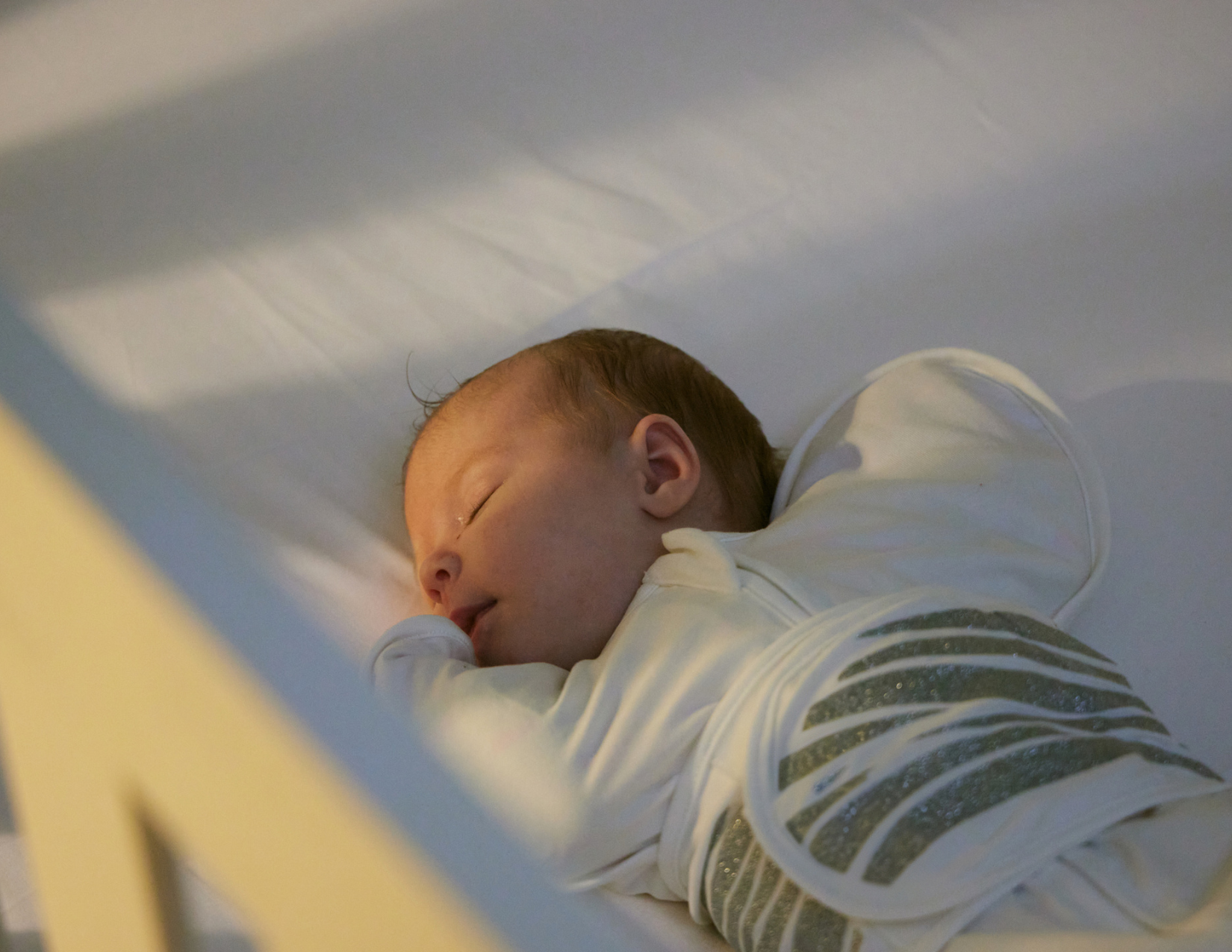 A baby sleeps peacefully in a crib, wrapped in a white swaddle with gray stripes, resting on a white sheet.