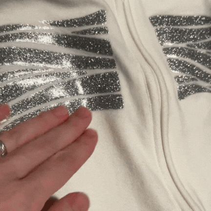 A hand with a ring rubs silver glittery stripes on a white fabric, revealing that one stripe changes color from silver to rainbow as its touched.