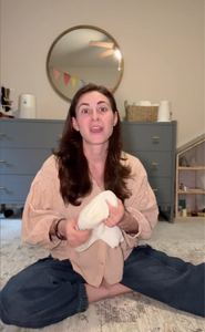 A woman with long brown hair sits cross-legged on the floor, holding a white towel. Behind her is a blue dresser, a mirror, and a dollhouse. The room has soft lighting and neutral decor.