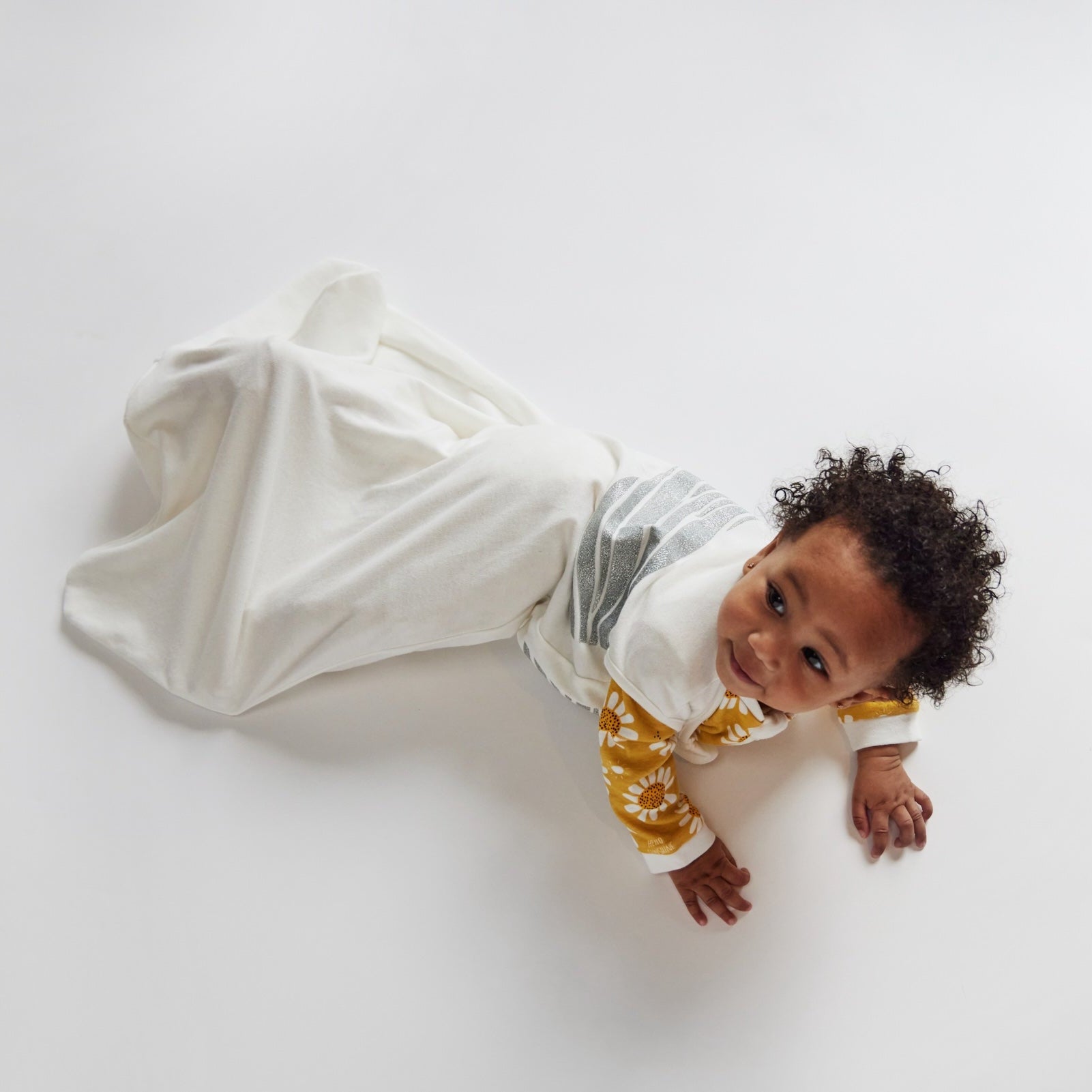 A baby dressed in items from the Baby Shower Bundle | 0 - 6 months, featuring a yellow floral top and a cozy white sleep sack, lies on a white surface, gazing upward with a curious expression.