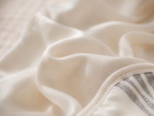 Close-up of soft cream-colored fabric with gentle folds and a small gray striped detail near the lower right edge.